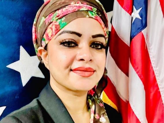 Woman in a floral headscarf and dark blazer standing in front of American flags and a patriotic background with stars.